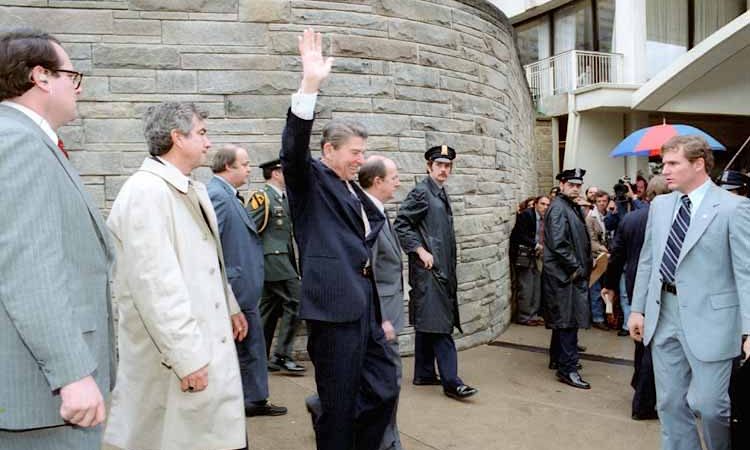 O presidente Ronald Reagan acena aos jornalistas à saída do Hotel Hilton. À esquerda, de gabardina branca, o agente Jerry Parr.