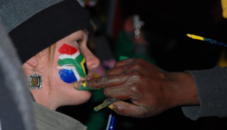 África do Sul, naão arco-íris: homem negro pinta a bandeira na cara de jovem branca