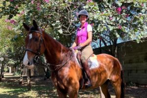 Cavaleira Luciana Diniz em cima de um cavalo castanho e a sorrir, com flores cor-de-rosa por trás.