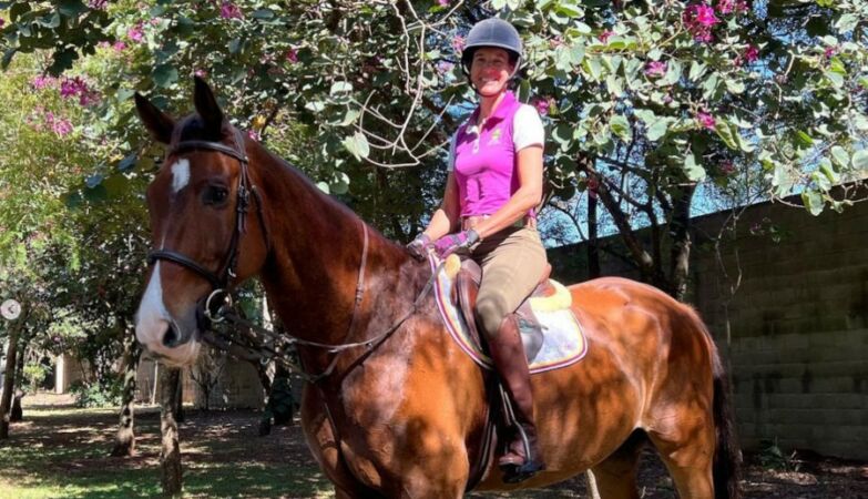 Cavaleira Luciana Diniz em cima de um cavalo castanho e a sorrir, com flores cor-de-rosa por trás.