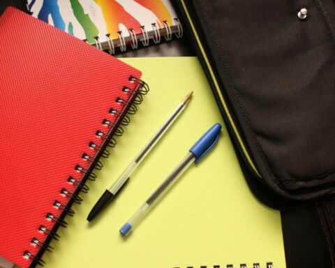 Caderno vermelho, duas canetas sobre caderno amarelo e estojo preto em artigo sobre o preço do material escolar.