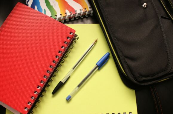 Caderno vermelho, duas canetas sobre caderno amarelo e estojo preto em artigo sobre o preço do material escolar.