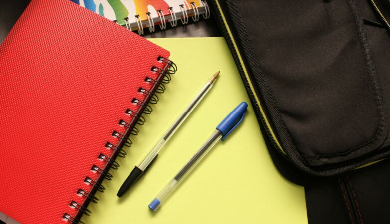 Caderno vermelho, duas canetas sobre caderno amarelo e estojo preto em artigo sobre o preço do material escolar.