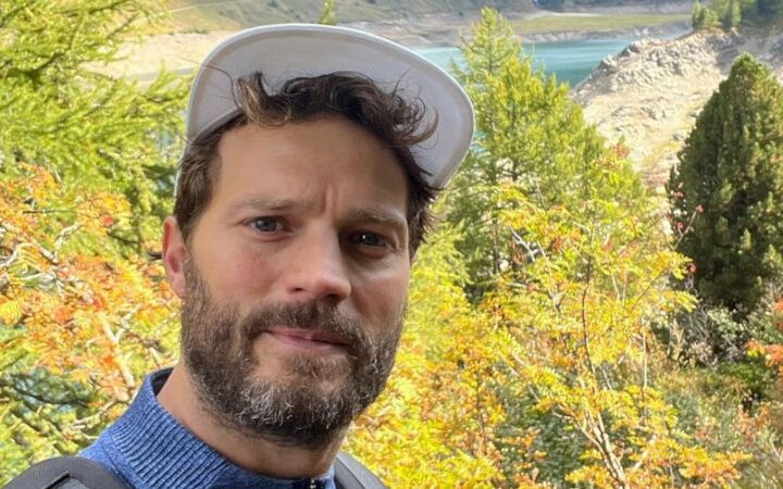 O actor Jamie Dornan de boné branco e barba grisalha a tirar uma selfie na montanha.
