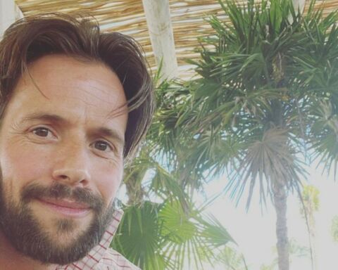 Actor Christian Oliver a sorrir com barba e palmeiras atrás.