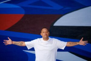 O músico Carlão de braços abertos, com tatuagens nos braços e t-shirt branca num fundo colorido em tons de azul.