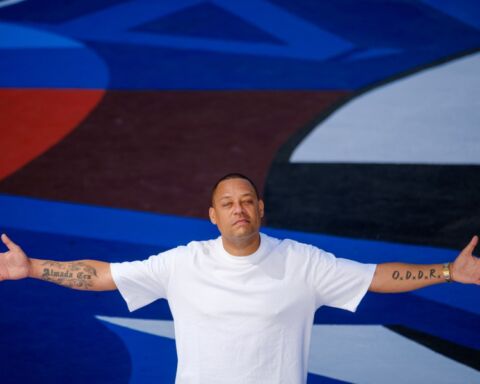 O músico Carlão de braços abertos, com tatuagens nos braços e t-shirt branca num fundo colorido em tons de azul.