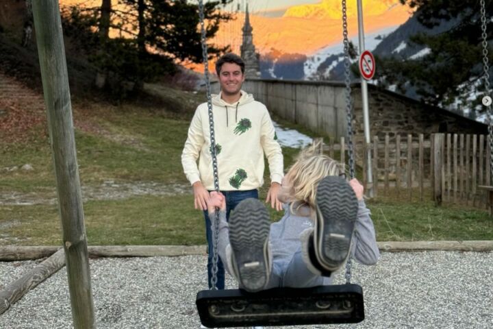 Gabriel Attal sorridente, de jeans e camisola branca, a empurrar o baloiço de uma criança loira num jardim.